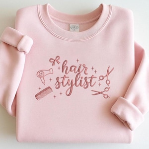 May include: A pale pink sweatshirt featuring the embroidered text "hair stylist" in a cursive script, accompanied by illustrations of a hairdryer, comb, and scissors. The design is in a matching pink colour.