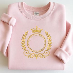 以下が含まれることがあります： 金色の刺繍デザインが施された、薄いピンク色のスウェットシャツ。デザインは、円の上に王冠、月桂樹の葉と装飾的な渦巻きで囲まれています。スウェットシャツはクルーネックと袖口が付いています。