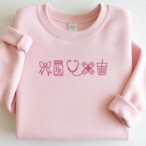 May include: A light pink sweatshirt with embroidered medical-themed icons in a darker pink. The icons include a bow, prescription bottle, stethoscope, bandage, and a drink with a straw. The sweatshirt has a crew neck and long sleeves.