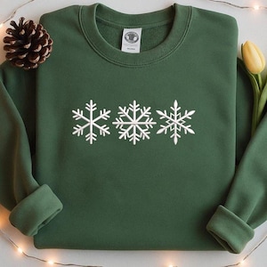 May include: A dark green sweatshirt featuring three white snowflakes across the chest. The sweatshirt has a crew neck and long sleeves. A pinecone and a yellow tulip are in the background.