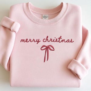 May include: A pale pink sweatshirt featuring the embroidered phrase "merry christmas" in a cursive script, accompanied by a red bow illustration. The garment has a crew neck and long sleeves with turned-up cuffs. A Gildan brand label is visible.