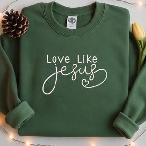 May include: A dark green sweatshirt with the words "Love Like Jesus" in white script. The word "Jesus" is in a cursive font with a heart symbol. The sweatshirt has a crew neck and long sleeves.