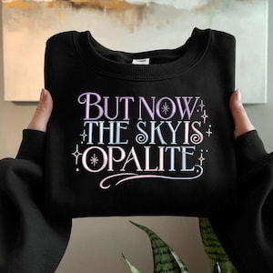 May include: Black crew neck sweatshirt with the text "BUT NOW THE SKY IS OPALITE" in a pastel color scheme. The text is in a decorative font with star and swirl accents. The sweatshirt is being held by a person.