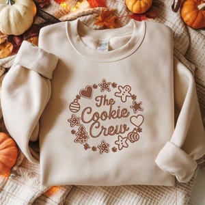 Op de afbeelding: Een beige sweatshirt met de tekst "The Cookie Crew" in een bruin cirkelvormig ontwerp. Het ontwerp bevat peperkoekmannen, sterren, harten en een kerstboom. De sweatshirt heeft lange mouwen en een ronde hals.