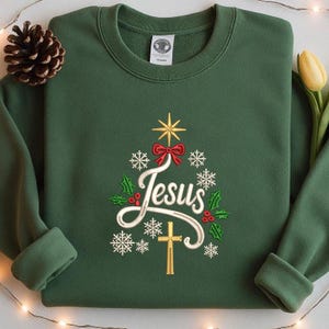 May include: Green sweatshirt with a Christmas tree design embroidered with the word "Jesus" in white script. The tree features a gold star, red bow, snowflakes, holly, and a gold cross. A pine cone and a yellow tulip are in the background.