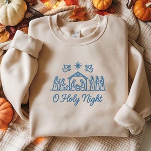 May include: A beige sweatshirt with blue embroidered design. The design features a nativity scene with the text "O Holy Night". The sweatshirt has long sleeves and a crew neck.