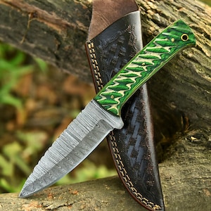 May include: A Damascus steel knife with a serrated blade and a green and white handle. The knife is paired with a black leather sheath with a basket weave pattern. The knife is resting on a piece of wood.