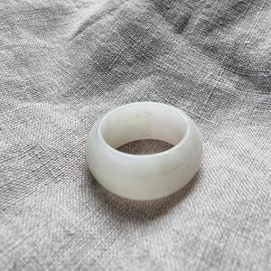 Puede incluir: Un anillo de banda ancha y lisa fabricado a partir de una pieza sólida de piedra translúcida de color blanco pálido. El anillo presenta una superficie exterior redondeada y pulida con sutiles variaciones de opacidad y un perfil circular.