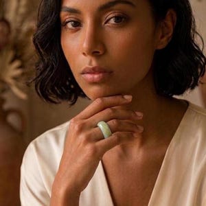 Puede incluir: Un anillo de banda de jade verde pálido liso con un acabado pulido. El anillo presenta un diseño circular ancho y sólido con un grosor constante, mostrando el color verde claro natural y las variaciones sutiles en la textura de la piedra.