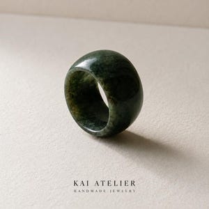 May include: A smooth, polished dark green stone ring with a wide, rounded band. The surface of the ring displays natural mottled patterns and variations in colour. Below the ring, the text KAI ATELIER HANDMADE JEWELRY is printed in black.