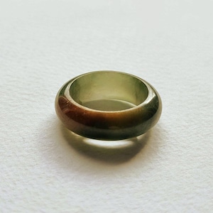 Op de afbeelding: Een gladde, gepolijste bandring met een kleurverloop van doorschijnend lichtgroen naar een diepe, aardse bruine kleur. De ring heeft een afgerond, dik profiel met een glanzende afwerking die het licht reflecteert over het gebogen oppervlak.