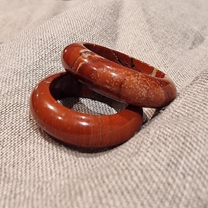 Peut inclure: Deux bracelets joncs circulaires polis en pierre brun rougeâtre. La surface des bracelets présente des variations naturelles, notamment des inclusions blanches tachetées et de subtils motifs linéaires dans le matériau opaque, lisse et brillant.