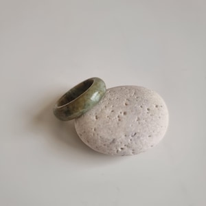 May include: A jade ring in shades of green sits on a light grey, textured stone. The ring is circular and appears to be carved from a single piece of jade. The stone has a smooth, rounded shape with small holes.