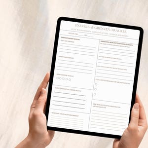 以下が含まれることがあります： ドイツ語の「エネルギーと境界線トラッカー」が表示された黒いタブレット。エネルギーレベル、意思決定、フォーカスに関するセクションがあります。タブレットは、ニュートラルな背景に対して手で持たれています。