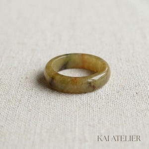 Op de afbeelding: Een gladde, gepolijste ringband gemaakt van natuursteen met een gevarieerd uiterlijk. De ring heeft aardse tinten van olijfgroen, goudgeel en bruin, met een glanzende afwerking die de unieke minerale insluitingen in de steen benadrukt.