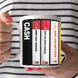 Peut inclure: Mug en c&eacute;ramique blanche avec des pochettes d'albums de Johnny Cash. La tasse affiche des titres d'albums tels que "Unchained", "American Recordings" et "Man in Black". La tasse est tenue par une personne avec du vernis &agrave; ongles rouge.