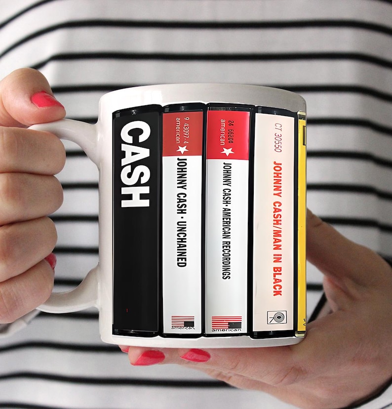 Peut inclure: Mug en c&eacute;ramique blanche avec des pochettes d'albums de Johnny Cash. La tasse affiche des titres d'albums tels que "Unchained", "American Recordings" et "Man in Black". La tasse est tenue par une personne avec du vernis &agrave; ongles rouge.