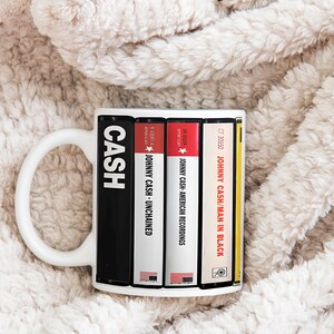 Peut inclure: Mug en c&eacute;ramique blanche avec un design inspir&eacute; des pochettes d'albums de Johnny Cash. La tasse affiche le mot "CASH" en lettres blanches en gras, ainsi que des titres d'albums comme "UNCHAINED" et "MAN IN BLACK". La tasse est pos&eacute;e sur un fond doux et textur&eacute;.