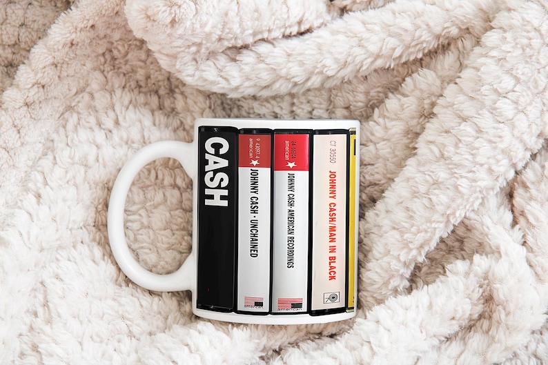 Peut inclure: Mug en c&eacute;ramique blanche avec un design inspir&eacute; des pochettes d'albums de Johnny Cash. La tasse affiche le mot "CASH" en lettres blanches en gras, ainsi que des titres d'albums comme "UNCHAINED" et "MAN IN BLACK". La tasse est pos&eacute;e sur un fond doux et textur&eacute;.