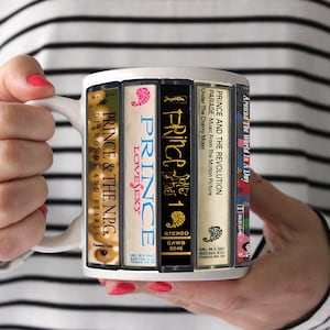 May include: White ceramic mug featuring album art from Prince's discography, including "Diamonds and Pearls," "Lovesexy," and "Parade." The mug is held by a person wearing a black and white striped shirt. A unique gift for music fans.