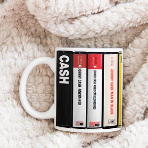 Peut inclure: Mug en c&eacute;ramique blanche avec un design inspir&eacute; des pochettes d'albums de Johnny Cash. Le mug affiche le mot "CASH" et des titres d'albums comme "Unchained" et "Man in Black" en pile verticale. La tasse a une anse blanche.
