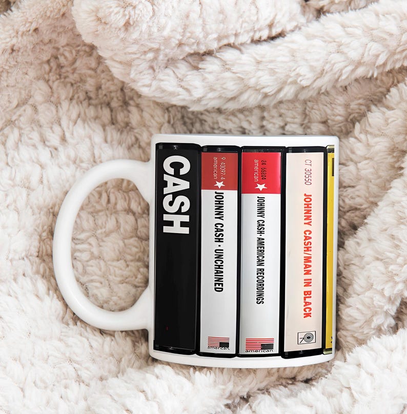 Peut inclure: Mug en c&eacute;ramique blanche avec un design inspir&eacute; des pochettes d'albums de Johnny Cash. Le mug affiche le mot "CASH" et des titres d'albums comme "Unchained" et "Man in Black" en pile verticale. La tasse a une anse blanche.