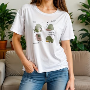 Peut inclure: T-shirt blanc avec un motif de cycle de vie de dinosaure : manger, dormir, cartes perforées et répéter. Le design comprend un ordinateur, un dinosaure endormi et un dinosaure mangeant. Vêtement décontracté.