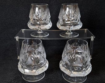 May include: Four clear crystal glasses with intricate etched designs. The glasses have a short, wide bowl and a stemmed base. They are arranged on a clear acrylic shelf against a black background. The cut crystal pattern features geometric and leaf-like shapes.