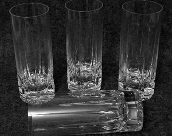 May include: A set of four clear glass tumblers featuring a tall cylindrical shape with vertical cut patterns extending from the heavy, faceted base. Three of the glasses stand upright, while one glass is positioned horizontally in the foreground.