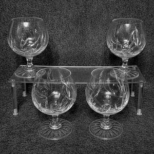 May include: A set of four clear glass snifters featuring a cut glass pattern of swirling lines on the bowls and a radial sunburst design on the circular bases. The glasses are arranged with two on a clear acrylic riser and two in front.
