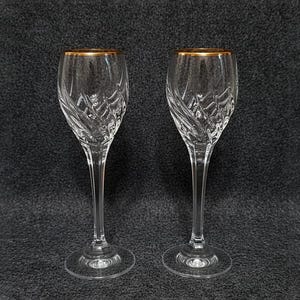 Può includere: Due calici di cristallo trasparente con bordi dorati. I calici hanno un gambo alto e sottile e un motivo a diamante decorativo inciso nella coppa. Su sfondo grigio scuro.