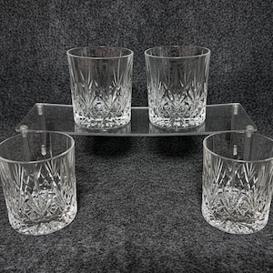 May include: Four clear, cut-glass tumblers with a diamond and vertical line pattern. Two tumblers are on a clear acrylic shelf, and two are on the surface below. The glasses are designed for serving beverages.