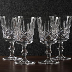 Könnte beinhalten: Vier klare Kristall-Weingläser mit aufwendigen Schliffmustern. Die Gläser haben ein klassisches Design mit einem Stiel und einer detaillierten Schale. Sie stehen auf einer dunklen Holzoberfläche vor einem dunklen strukturierten Hintergrund.