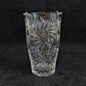 Könnte beinhalten: Eine transparente, geschliffene Glasvase mit Sternenmuster und geometrischem Muster. Die Vase hat einen gewellten Rand und eine zylindrische Form, etwa 20 cm hoch.