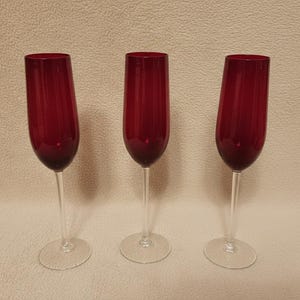 May include: Three red champagne flutes with clear stems and circular bases. The glasses are evenly spaced against a neutral background. The flutes have a classic, elegant design.