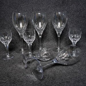 Puede incluir: Un conjunto de copas de vino de cristal transparente, algunas de pie y otras tumbadas. Las copas presentan un diseño detallado de cristal tallado y están dispuestas sobre una superficie gris oscuro. La imagen muestra una variedad de tamaños.