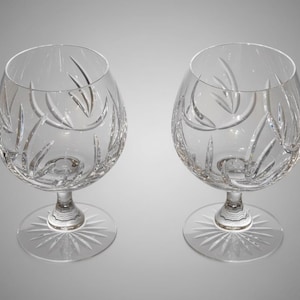 May include: Two clear crystal brandy glasses. Each glass has an oval bowl with etched leaf designs and a starburst pattern on the base. The stem is short and the bowl is wide.