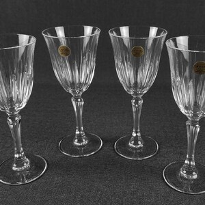 May include: A set of four clear crystal wine glasses featuring vertical fluted detailing on the bowls and stems. Each glass has a circular base and a small round gold-colored sticker on the side of the bowl with the text Crystal.