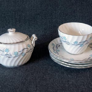 May include: A white porcelain tea set with a sugar bowl, a teacup, and saucers. The set features a spiral design with blue floral accents and silver trim. The set includes a sugar bowl with a lid and two handles.