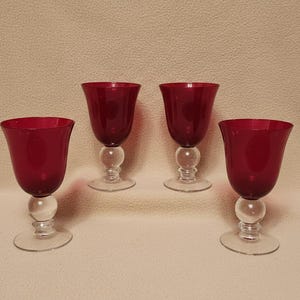 May include: Four ruby red wine glasses. Each glass features a rounded bowl, a clear, spherical stem, and a clear, circular base. The glasses are displayed against a neutral backdrop, highlighting their elegant design and colour.