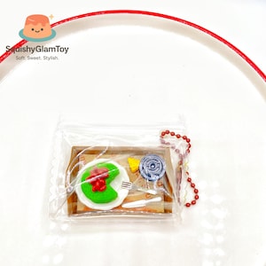 May include: A miniature food-themed charm set in a clear plastic bag. The set includes a green and red item, a yellow item, a blue item, and miniature silverware. The charm is on a white plate with a red rim. The text "SquishyGlamToy" is visible.