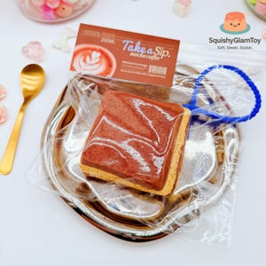 May include: A brown and tan mocha coffee squishy toy in a clear plastic bag, with a blue handle, on a silver tray. The packaging reads "Take a Sip. mocha coffee" with a coffee cup graphic. A gold spoon is visible.