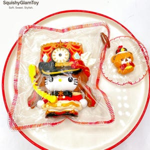 Könnte beinhalten: Ein weißer Teller mit rotem Rand enthält eine transparente Plastiktüte mit einer Hello Kitty-Figur in roter und schwarzer Uniform. Eine kleinere Tüte enthält eine Bärenfigur. Der Text "SquishyGlamToy" ist sichtbar.