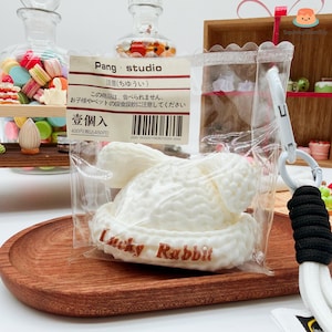 May include: A white, squishy rabbit-shaped toy in a clear plastic bag. The text "Lucky Rabbit" is printed in brown on the toy. The bag is attached to a white lanyard with a black accent. The background includes a wooden tray and decorative items.