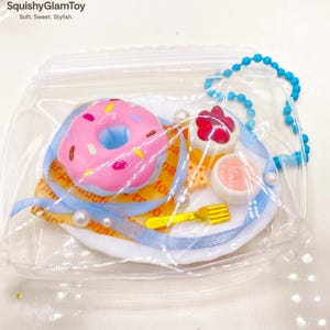 May include: A miniature food-themed keychain featuring a pink donut with sprinkles, a red ice cream cone, and a pink dessert on a white plate. The plate is decorated with a blue ribbon, faux pearls, and a gold fork. The keychain is in a clear plastic bag.