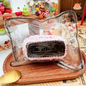 Könnte beinhalten: Eine transparente Plastiktüte mit schwarzem Reißverschluss enthält einen rosa-braunen Kuchen. Der Kuchen ist rechteckig mit einem dunkelbraunen Kern und rosa Zuckerguss. Weitere Miniaturdesserts und Süßigkeiten sind im Hintergrund zu sehen.