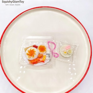 May include: A white ceramic plate with a red rim holds two small, clear plastic bags containing miniature food items. One bag features a plate with a miniature breakfast, and the other contains a miniature plate with eggs. The image includes the text "SquishyGlamToy. Soft. Sweet. Stylish."