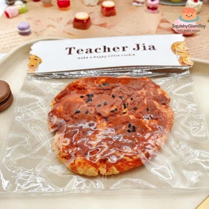 May include: A large, round, reddish-brown cookie with black sesame seeds, packaged in clear plastic. The label reads "Teacher Jia make a happy little cookie." The cookie is part of a collection of miniature food items.