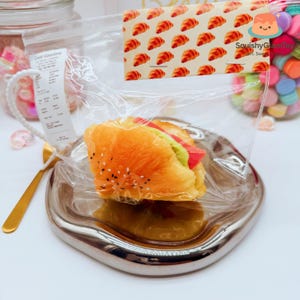 May include: A squishy toy shaped like a croissant sandwich, sealed in a clear plastic bag. The croissant is orange with sesame seeds, and the filling includes red and green elements. A tag with croissant pattern is attached to the bag.