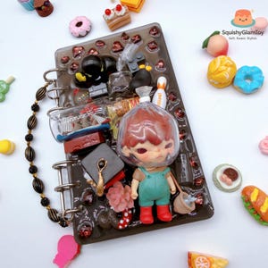 Puede incluir: Un diario decorativo con una variedad de dijes en miniatura, que incluyen una muñeca, alimentos y otros objetos pequeños. El diario es negro con una encuadernación en espiral y una cubierta transparente. El texto "SquishyGlamToy" es visible.